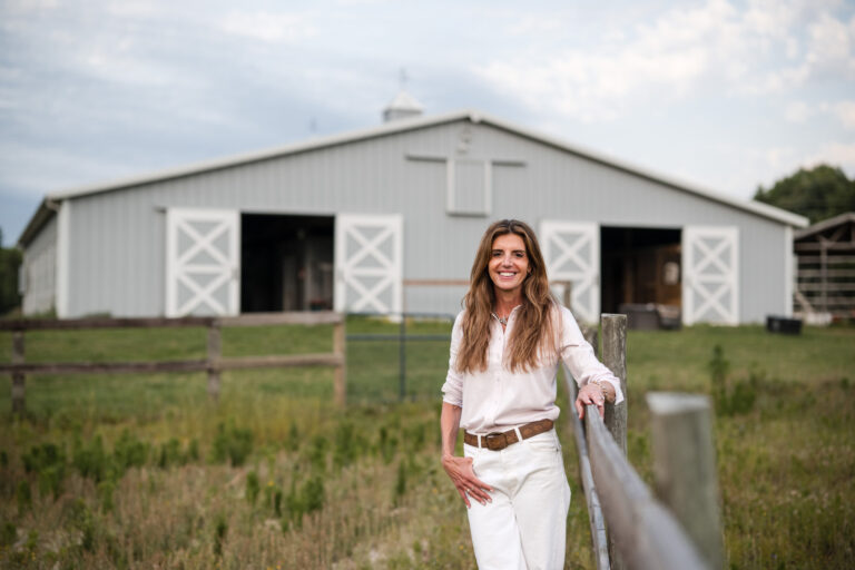 Smiling woman in white standing outdoors near a wooden fence with a large white barn in the background.